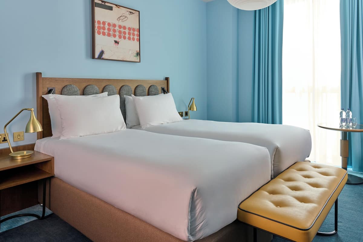 A hotel room with two beds, a bench, a desk with a lamp, and a framed picture on the wall.