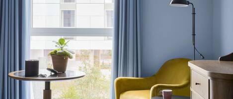A hotel room with a bed, a yellow armchair, a small round table with a lamp, a potted plant, and a view of buildings through a window with blue curtains.