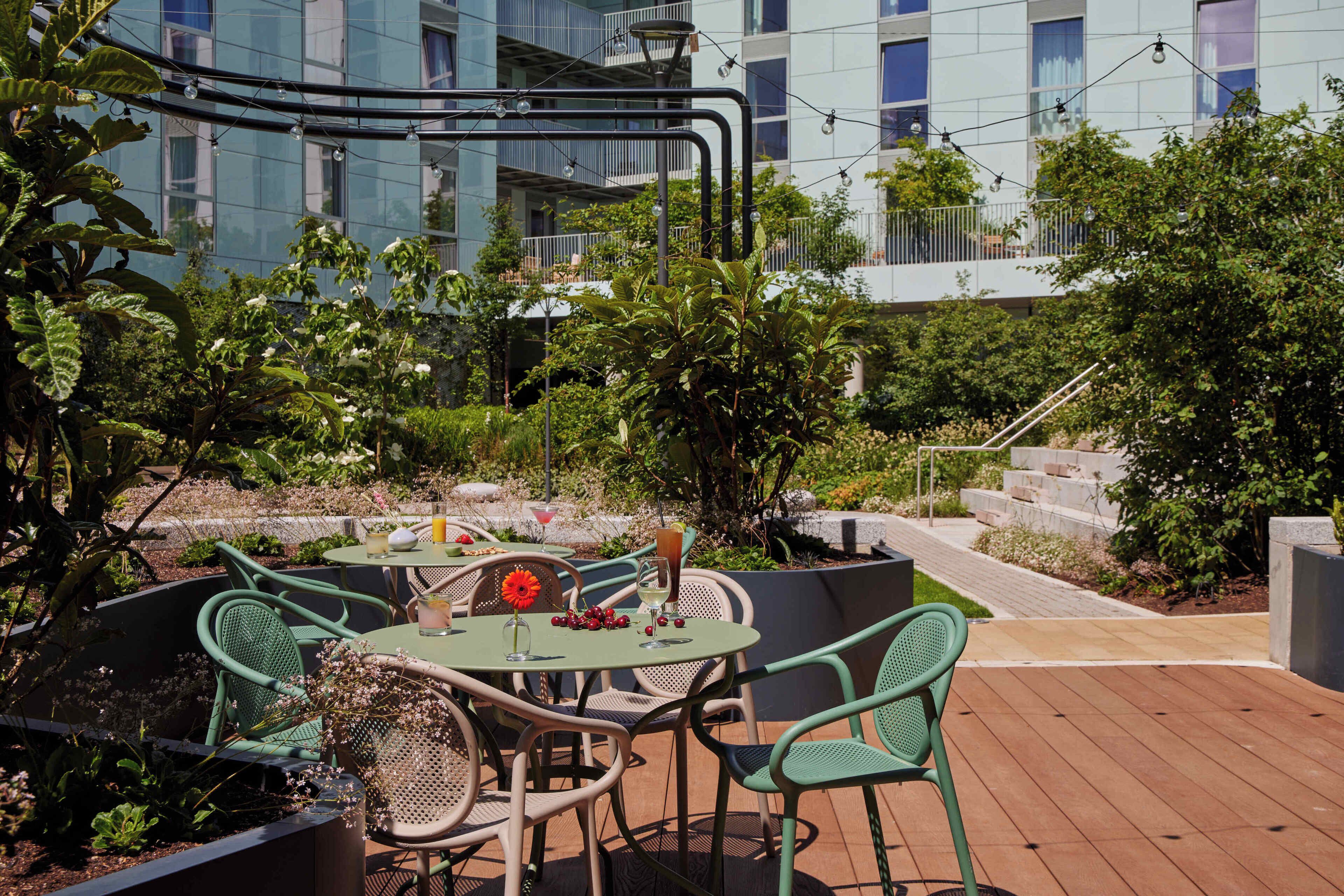 An outdoor patio with round tables and green chairs, surrounded by lush greenery and a modern building in the background.