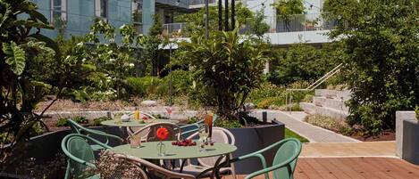 An outdoor patio with round tables and green chairs, surrounded by lush greenery and a modern building in the background.