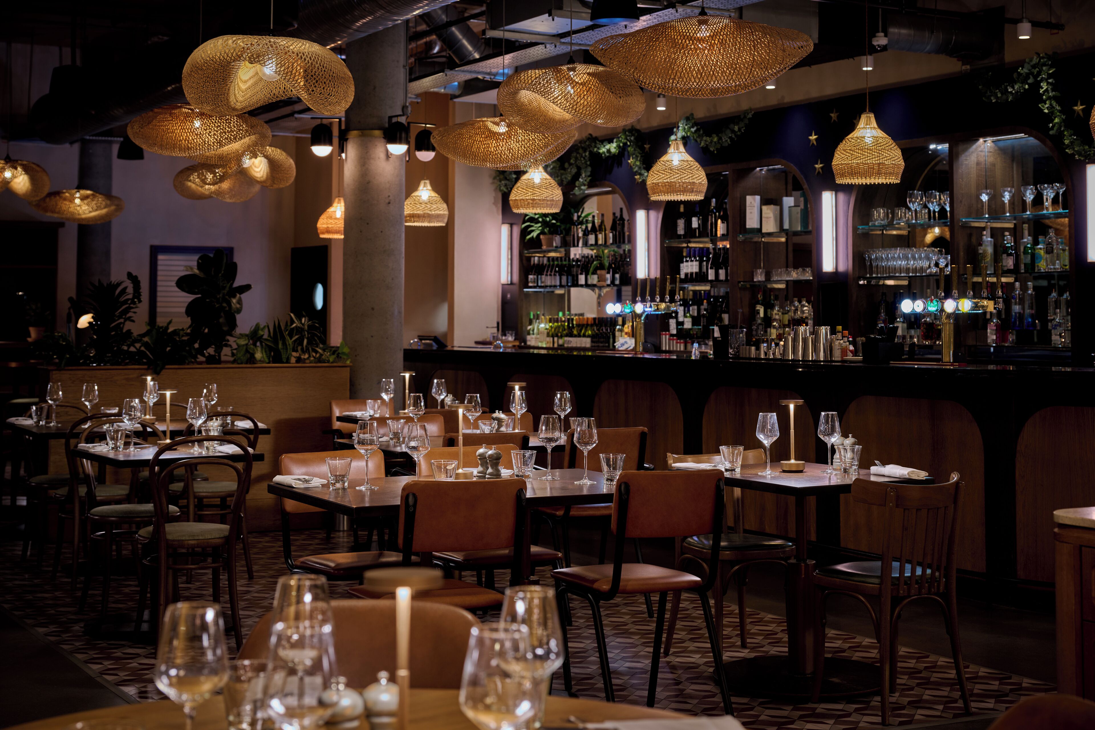 A restaurant with tables set for dining, hanging wicker chandeliers, and a bar area with shelves of bottles and glasses.
