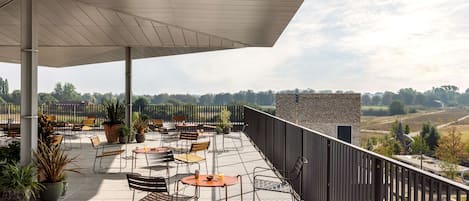 A covered outdoor seating area with tables and chairs, potted plants, and a view of the countryside.