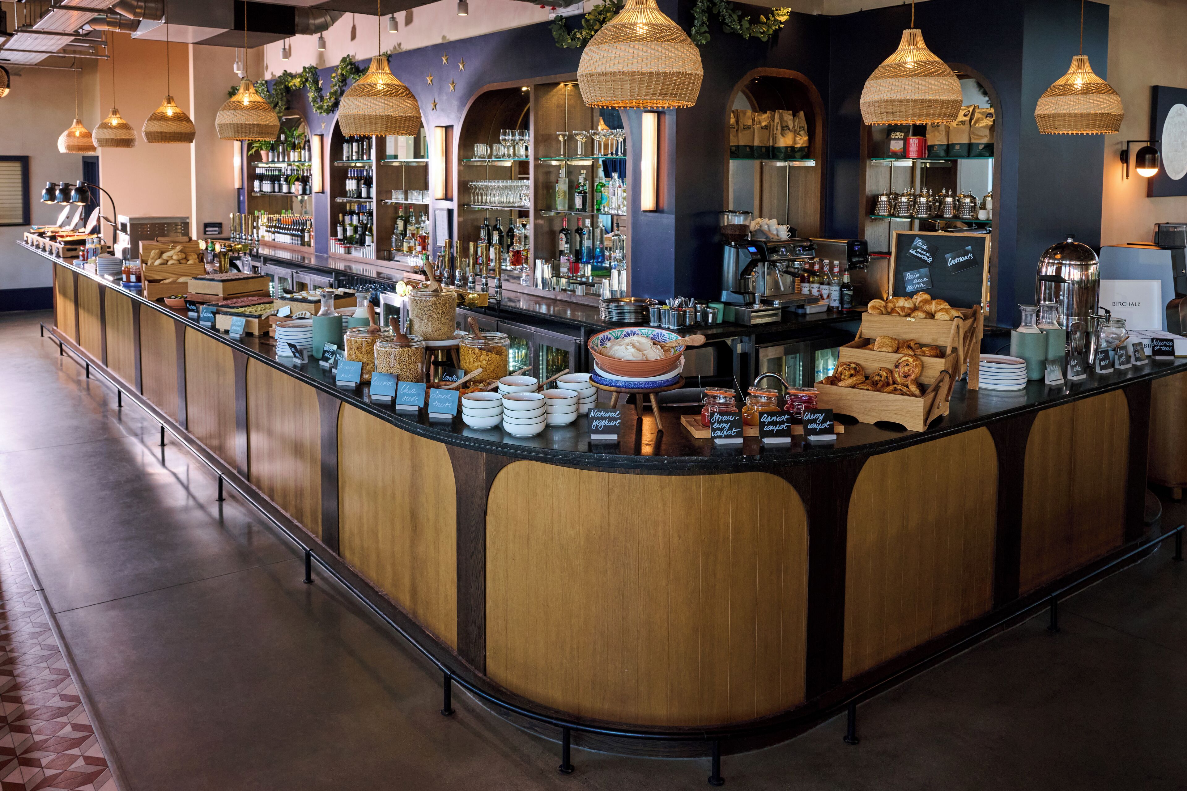 Zona de bar con mostrador curvo, iluminación colgante de mimbre y diferentes productos alimenticios y bebidas a la vista.