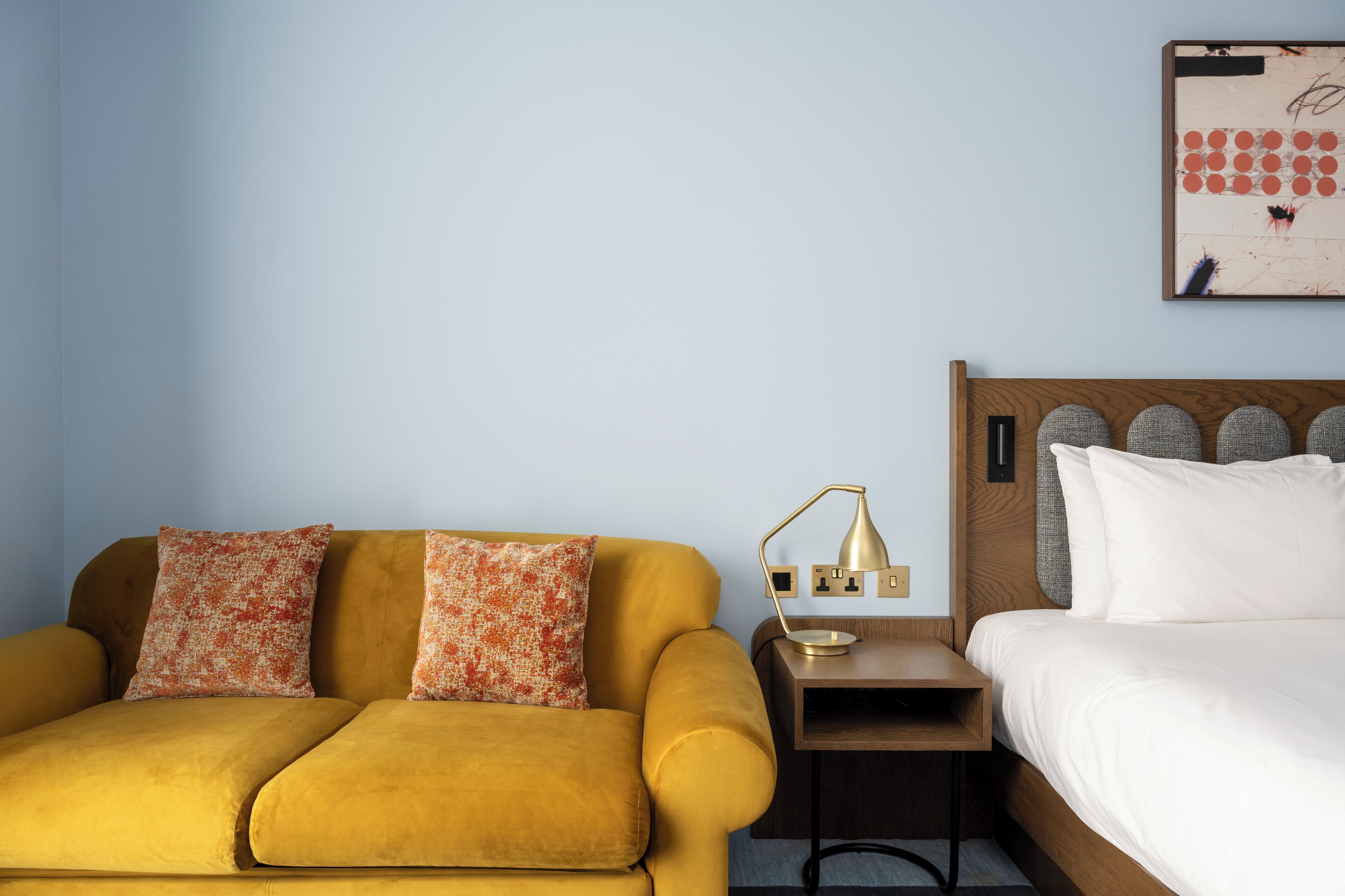 A room with a mustard-colored sofa, a small wooden nightstand with a lamp, and a bed with white bedding.
