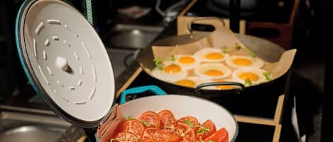 Een keuken met een blauwe pan met plakjes tomaat op een fornuis, een koekenpan met eieren en een pan deksel.