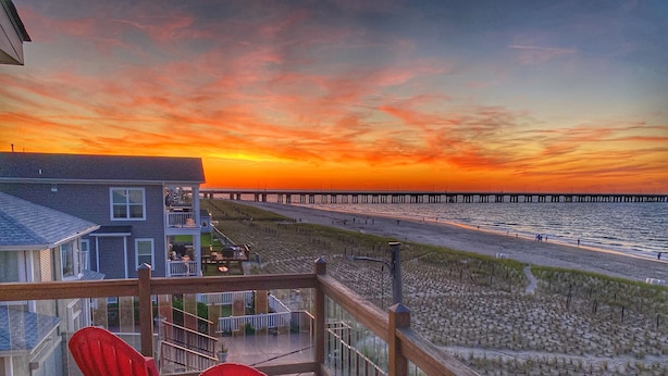 A wooden deck with red chairs overlooking a beach and a pier.