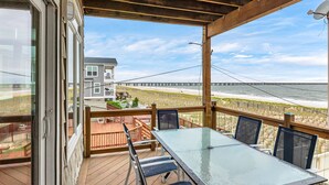 A covered outdoor deck with a glass table and chairs, overlooking a beach and a pier.
