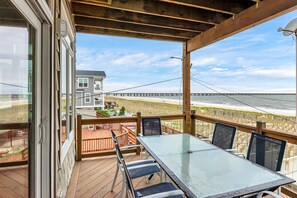 A covered outdoor deck with a glass table and chairs, overlooking a beach and a pier.