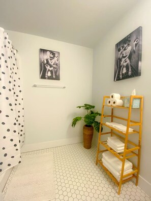 A bathroom with a shower curtain, two framed artworks, a potted plant, a wooden shelf with towels, and a hexagonal tiled floor. - The coolest loft in Cincinnati Area (Maineville)