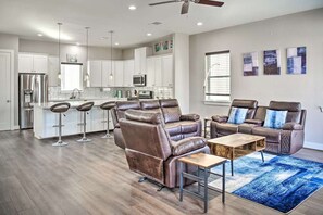 A modern living room with a leather recliner and a coffee table, a kitchen with white cabinets and stainless steel appliances, and a dining area with bar stools.