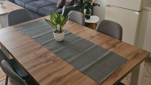 Ein Essbereich mit einem Holztisch, einer schwarz-weißen Tischdecke und einem Blumentopf. Im Hintergrund befindet sich ein graues Sofa und ein Kühlschrank.