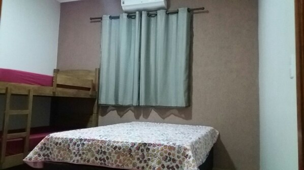 Une chambre avec un lit superposé, un lit simple recouvert d’une couvre-lit à motifs floraux, une fenêtre avec des rideaux et un climatiseur fixé au mur.