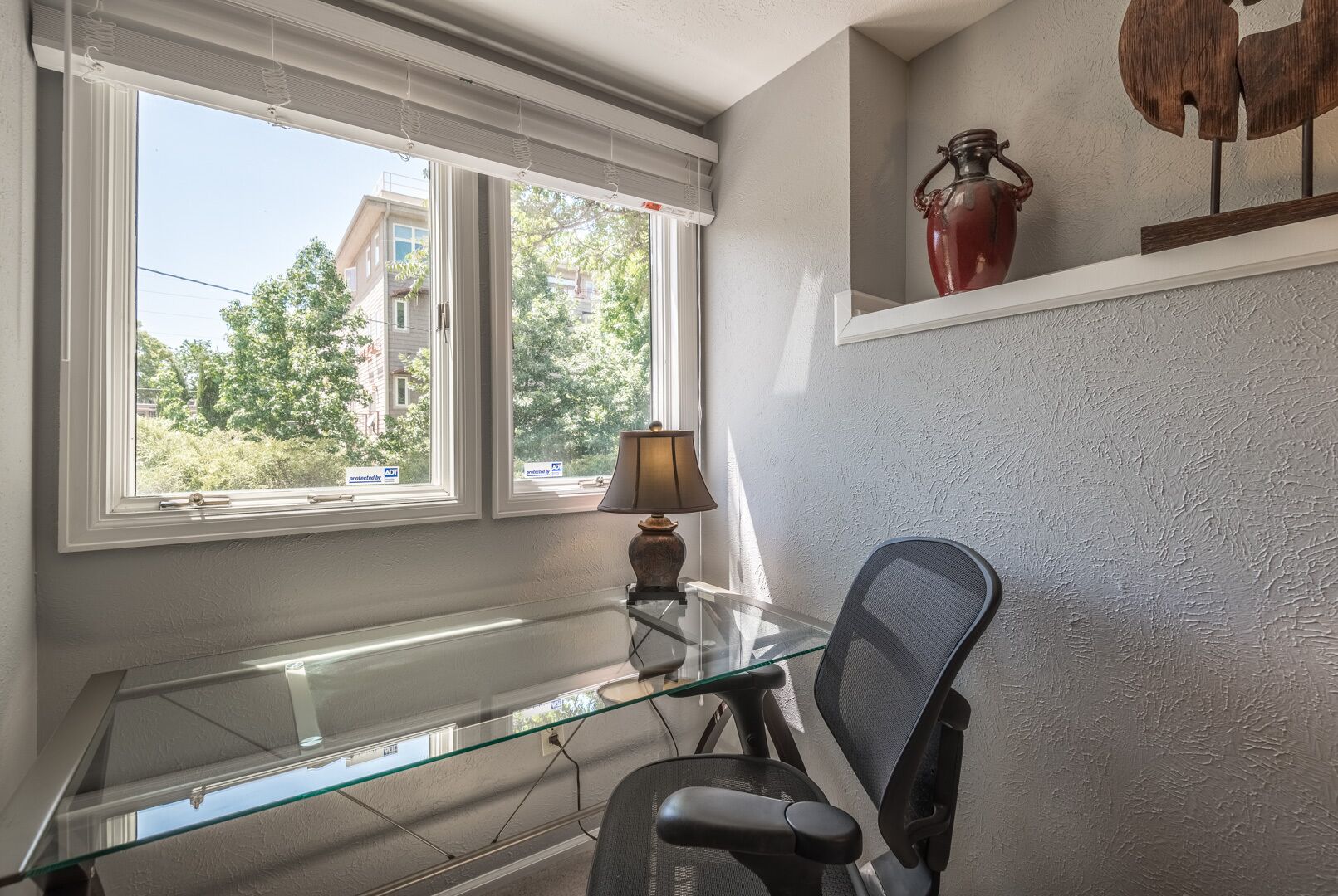 A room with a glass desk, a chair, a lamp, and a shelf with a vase.