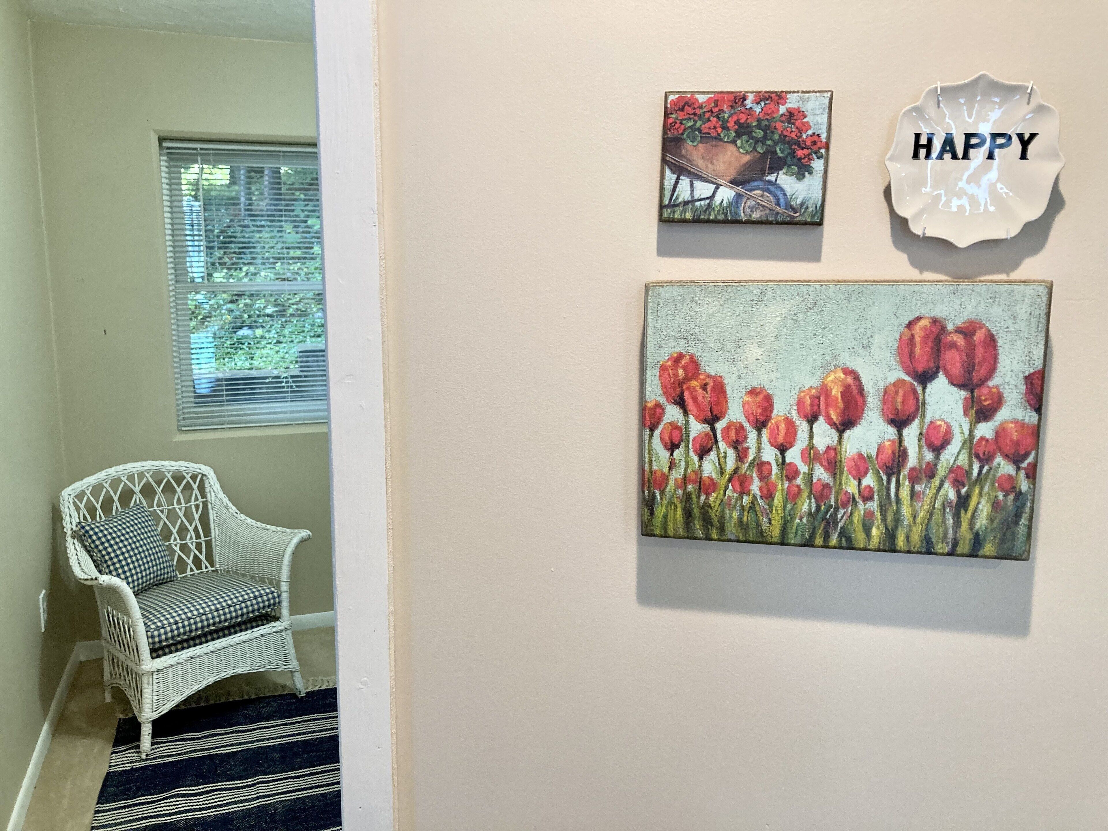 A hallway with a white wicker chair, a window with blinds, and two wall-mounted paintings of flowers.