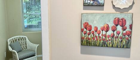 A hallway with a white wicker chair, a window with blinds, and two wall-mounted paintings of flowers.
