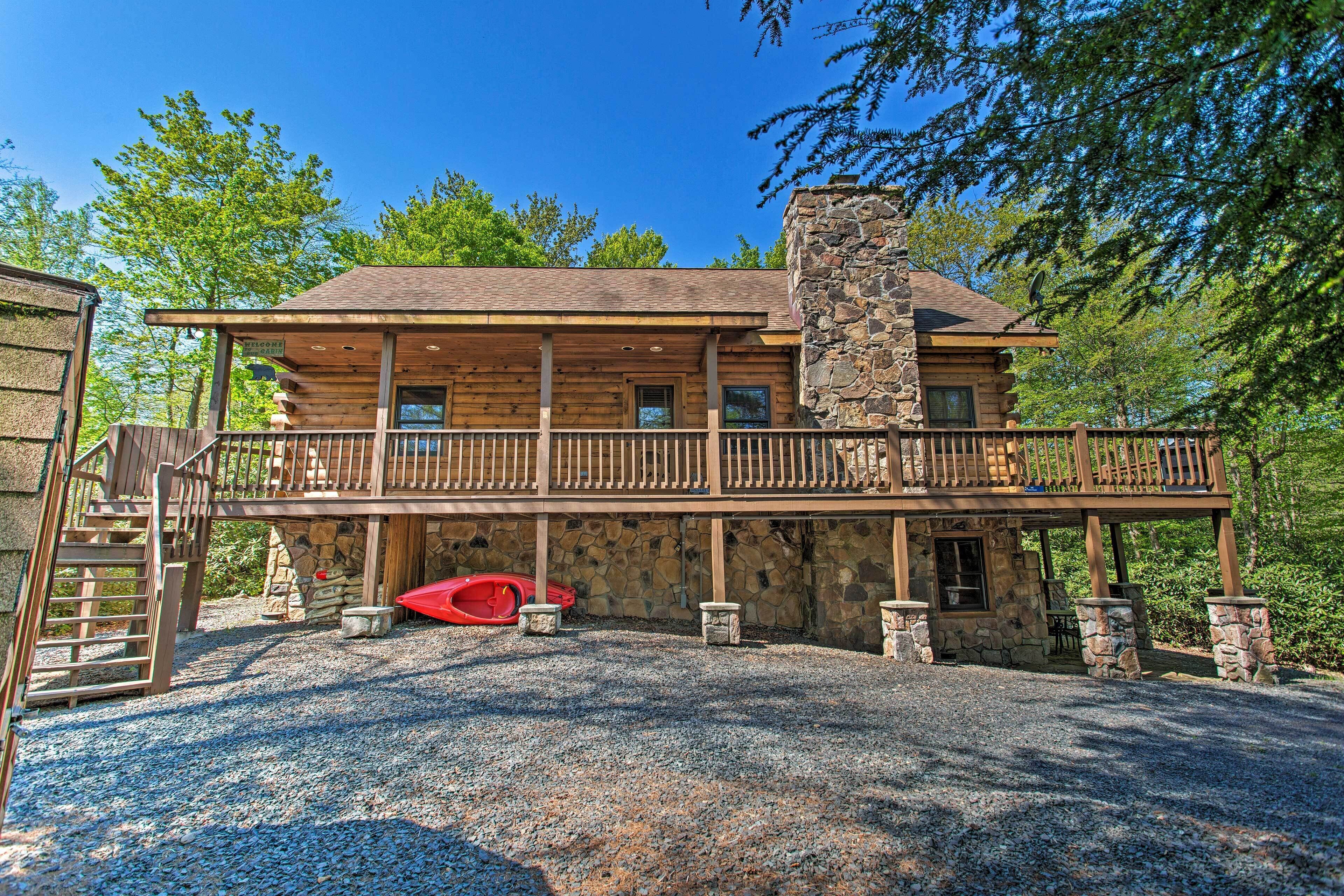 A log cabin with a stone chimney, a covered deck, and a gravel driveway.