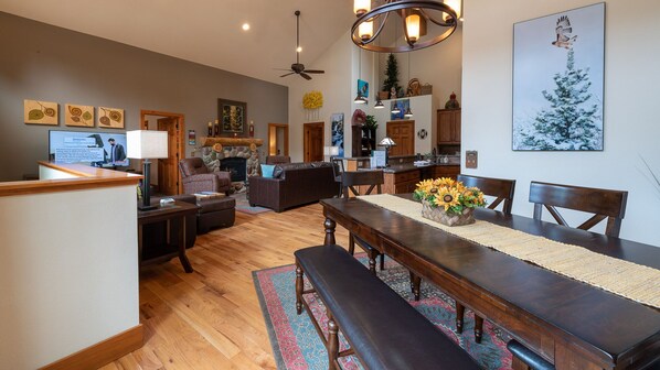 A spacious living room with a fireplace, a dining area with a long table and benches, and a kitchen area with wooden cabinets. - Family Gathering Home! Beautiful, Spacious, Clean! Next to Rocky Mtn N.P., YMCA (Estes Park)