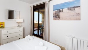 Habitación de hotel con cama, cómoda, ventilador de techo, ventana con cortinas y una pintura con vista al mar.
