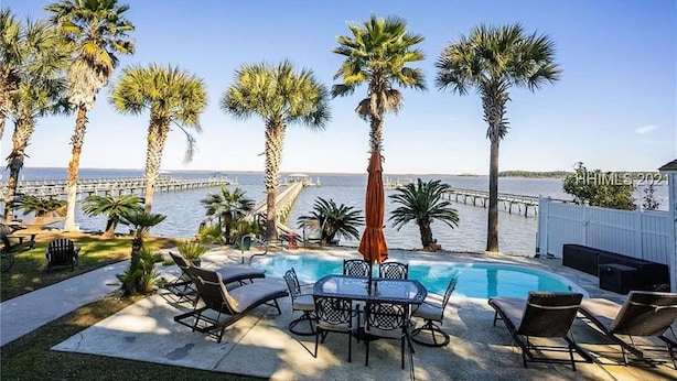 A pool area with lounge chairs and a table, palm trees, and a view of the ocean and a pier. - Dreamworthy, waterfront St. Helena home with pool and deep water dock. (St. Helena)