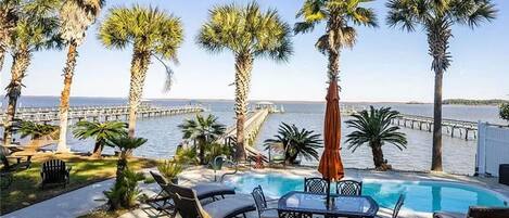 A pool area with lounge chairs and a table, palm trees, and a view of the ocean and a pier.
