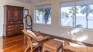 Quarto com armário de madeira, mobiliário de vime e amplas janelas com vista para palmeiras e um cais.