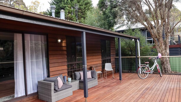 Une terrasse extérieure couverte avec un plancher en bois, un canapé en osier avec des coussins, un vélo et une porte vitrée donnant sur une pièce.