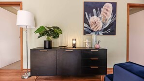Un salon moderne avec un parquet en bois foncé, un meuble noir avec des tiroirs, un lampadaire, une plante en pot, un tableau représentant des fleurs et un canapé bleu.