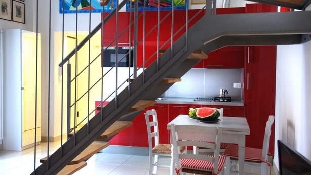 A modern kitchen with red cabinetry, a dining area with white chairs, and a staircase with a metal railing. - GetTheKey Boldrini Loft 2 (Bologna)