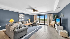 Un salon moderne avec un canapé gris, une table basse et un téléviseur posé sur un meuble blanc. On y trouve également un ventilateur de plafond, une grande fenêtre avec des rideaux et une vue sur la verdure à l’extérieur.