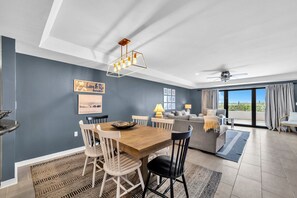 Un salon moderne avec un coin repas, comprenant une table à manger en bois, des chaises blanches et noires, un canapé et une grande fenêtre avec des rideaux.