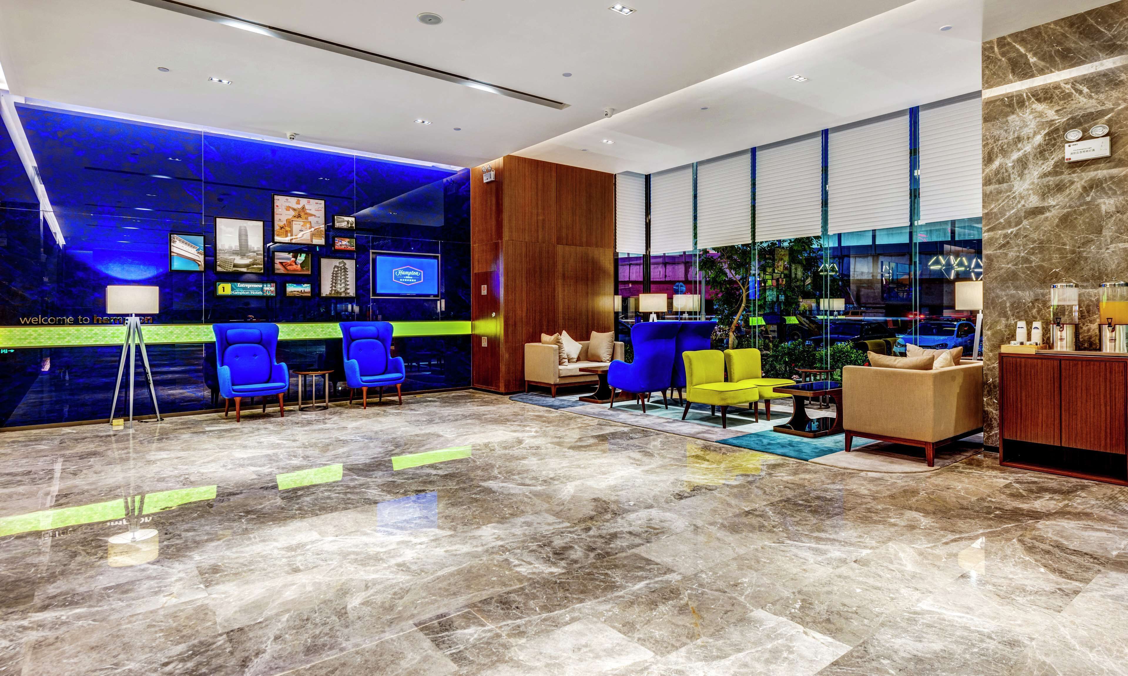 A modern hotel lobby with a blue and green color scheme, featuring a reception desk, seating area with blue and yellow chairs, and a large window offering a view of the city.
