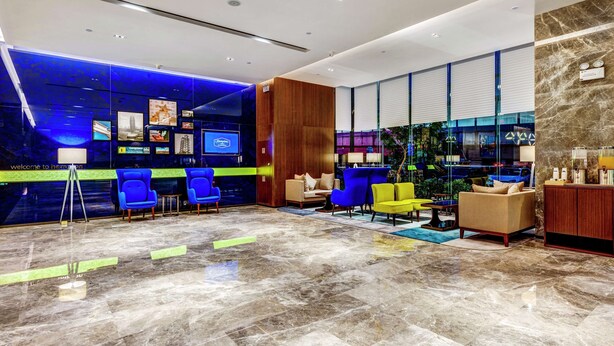 Un moderno vestíbulo de hotel con una paleta de colores azul y verde, que incluye una recepción, un área de descanso con sillas azules y amarillas y un amplio ventanal con vistas a la ciudad.
