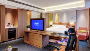 Habitación de hotel con cama, escritorio con computadora, televisor, silla y pequeña mesa con planta.