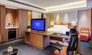 Habitación de hotel con cama, escritorio con computadora, televisor, silla y pequeña mesa con planta.
