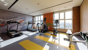 Un gimnasio moderno con cintas de correr, elípticas y una bicicleta estática. Amplios ventanales ofrecen vistas a la ciudad.