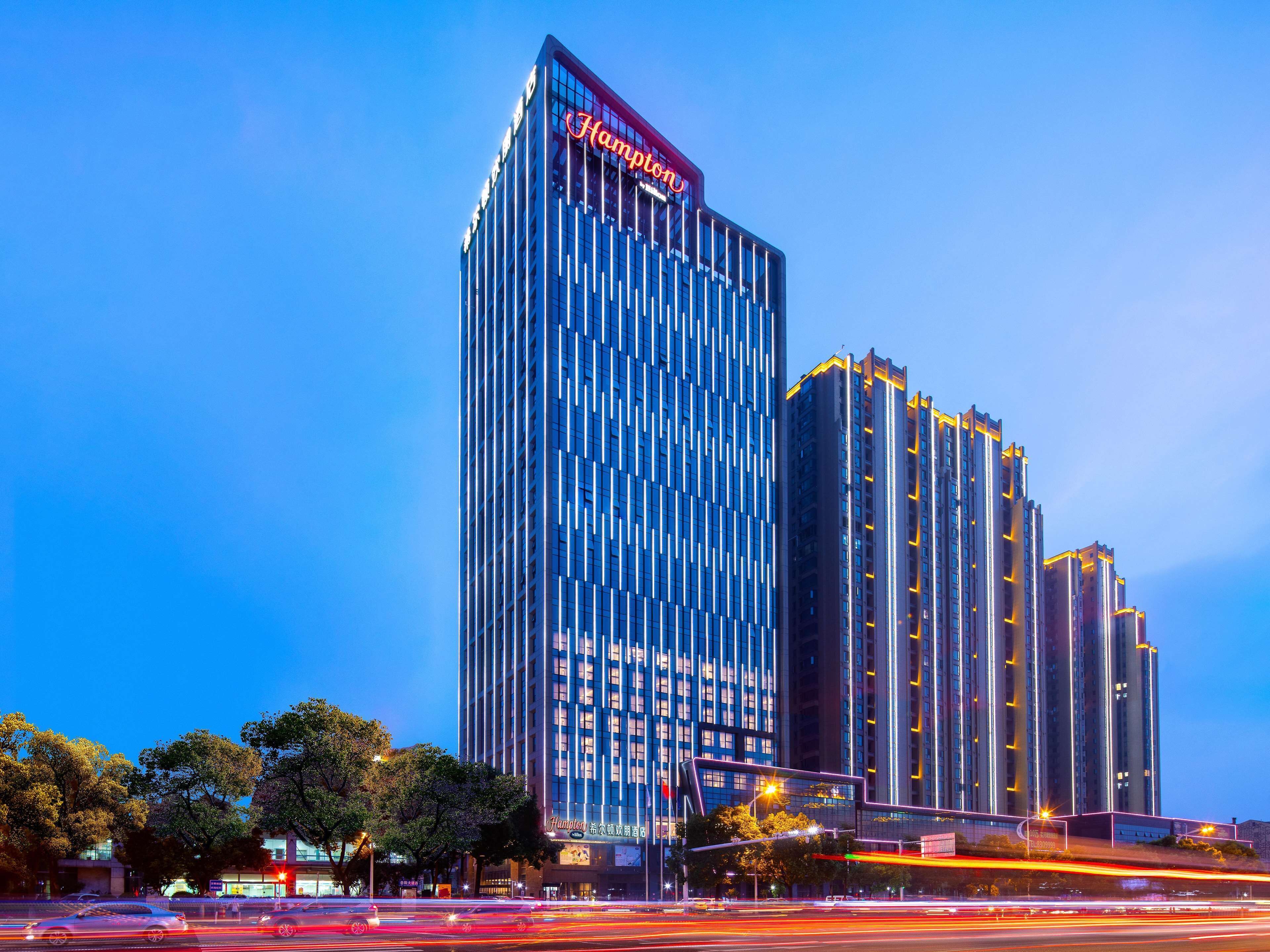 A high-rise hotel with a prominent 'Hampton' sign, featuring a modern architectural design with vertical lines and reflective glass windows.
