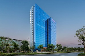 A modern high-rise building with a glass facade, surrounded by greenery and a clear sky.