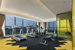 Ein modernes Fitnessstudio mit Laufbändern, Crosstrainern und einer Bank. Große Fenster bieten Blick auf die Stadt.