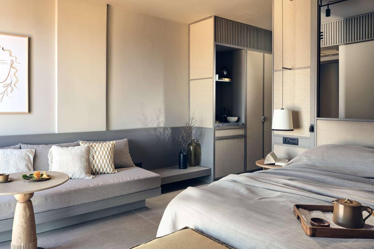 A modern hotel room with a sofa, a bed, a small table, a bookshelf, and a hanging light fixture.