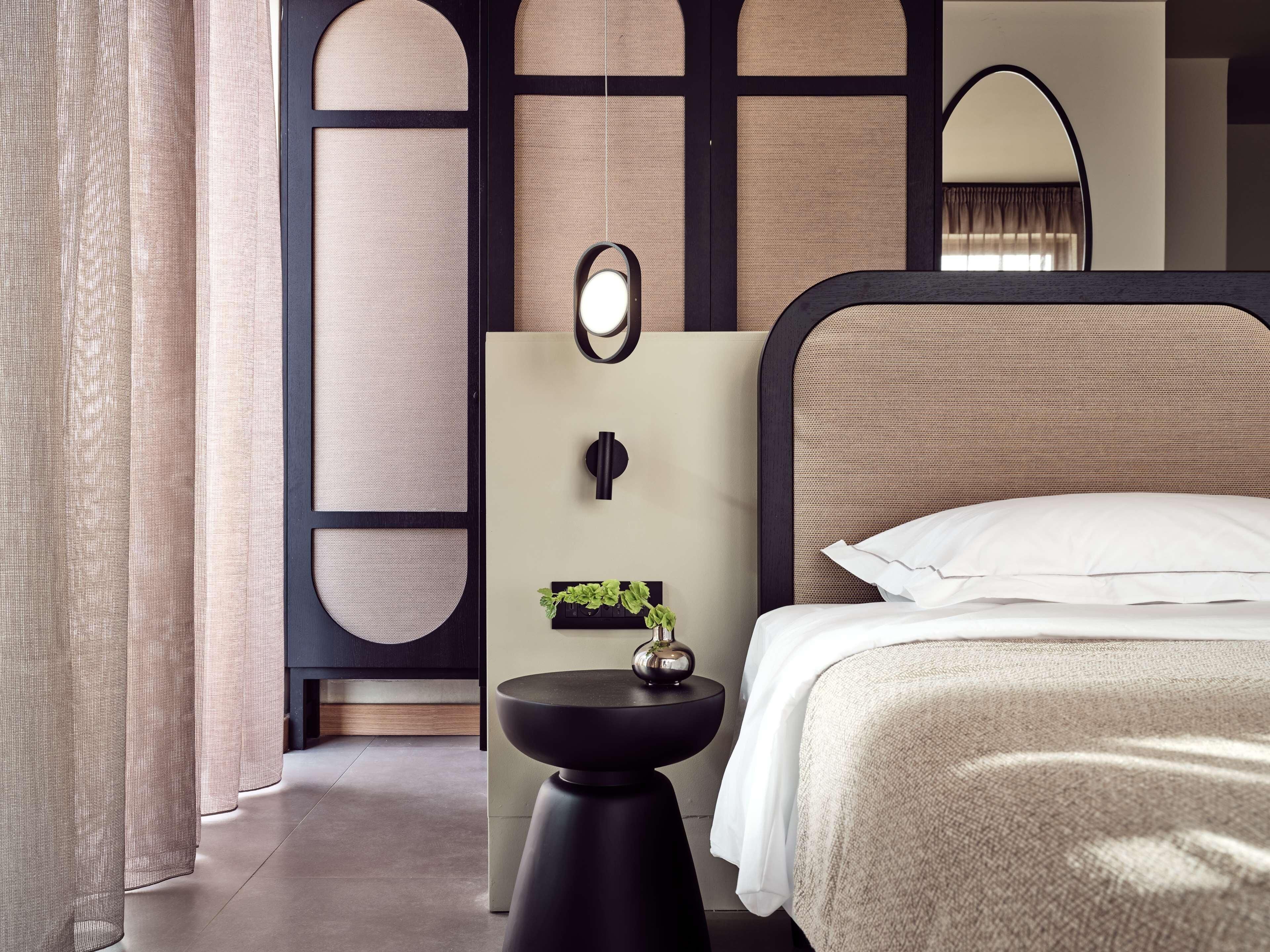 Une chambre d’hôtel moderne avec un lit, un miroir rond, une petite table d’appoint avec une plante et un panneau décoratif au mur.