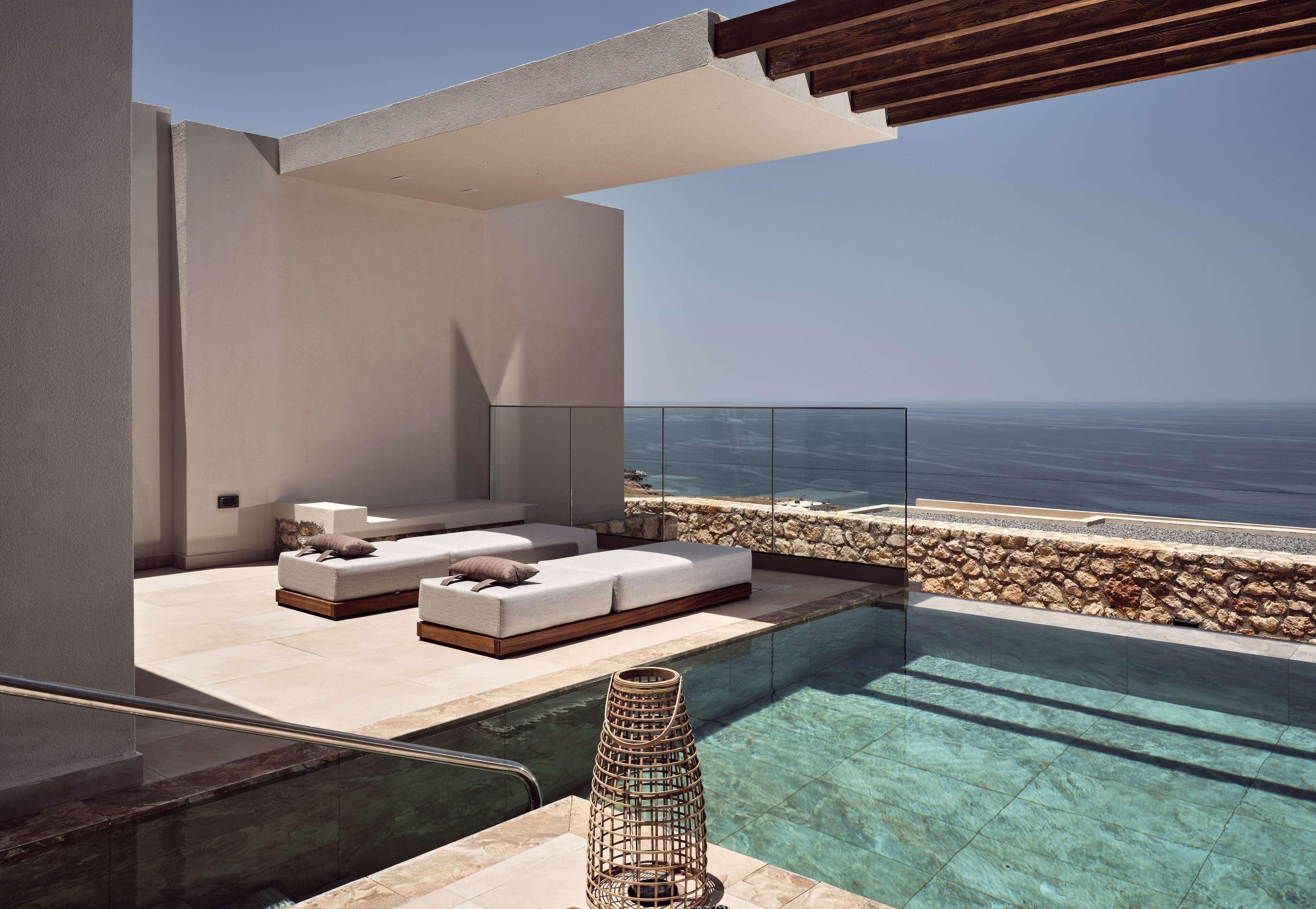 Une piscine à débordement moderne offrant une vue dégagée sur l’océan, un espace détente avec des coussins et un mur en pierre.