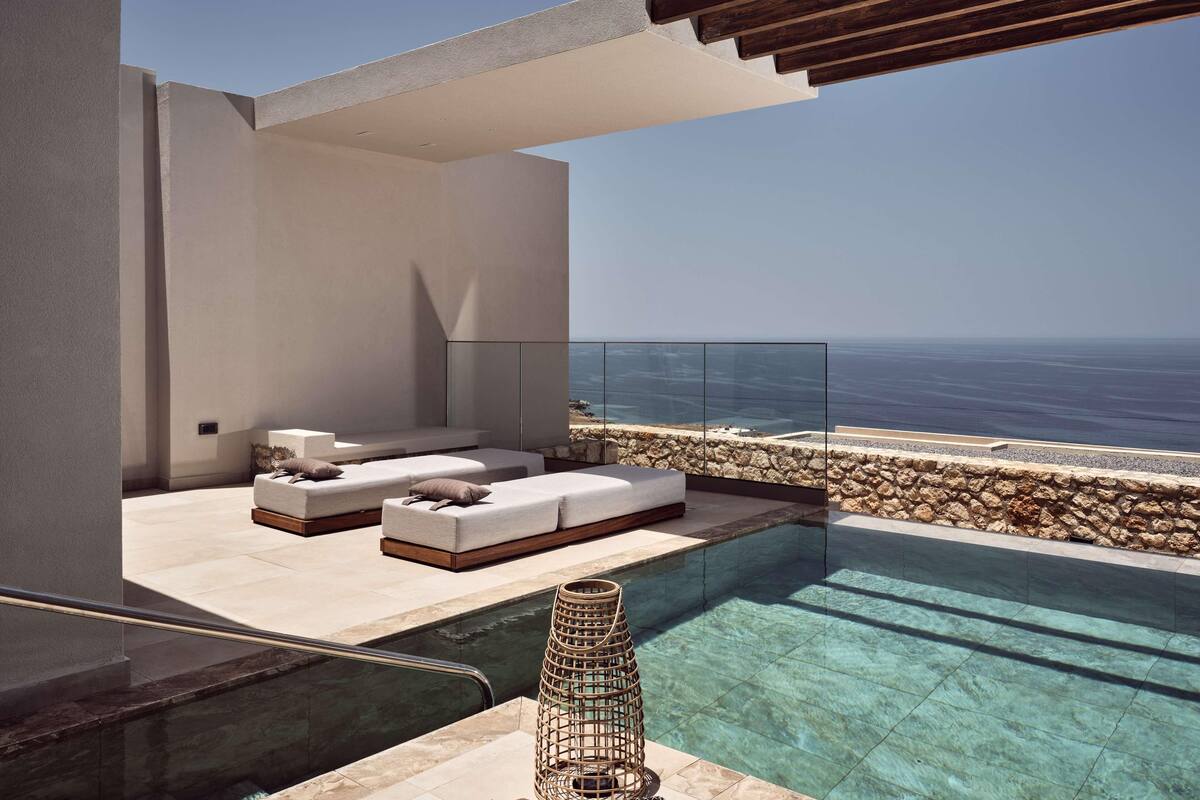 A modern infinity pool with a clear view of the ocean, a seating area with cushions, and a stone wall.