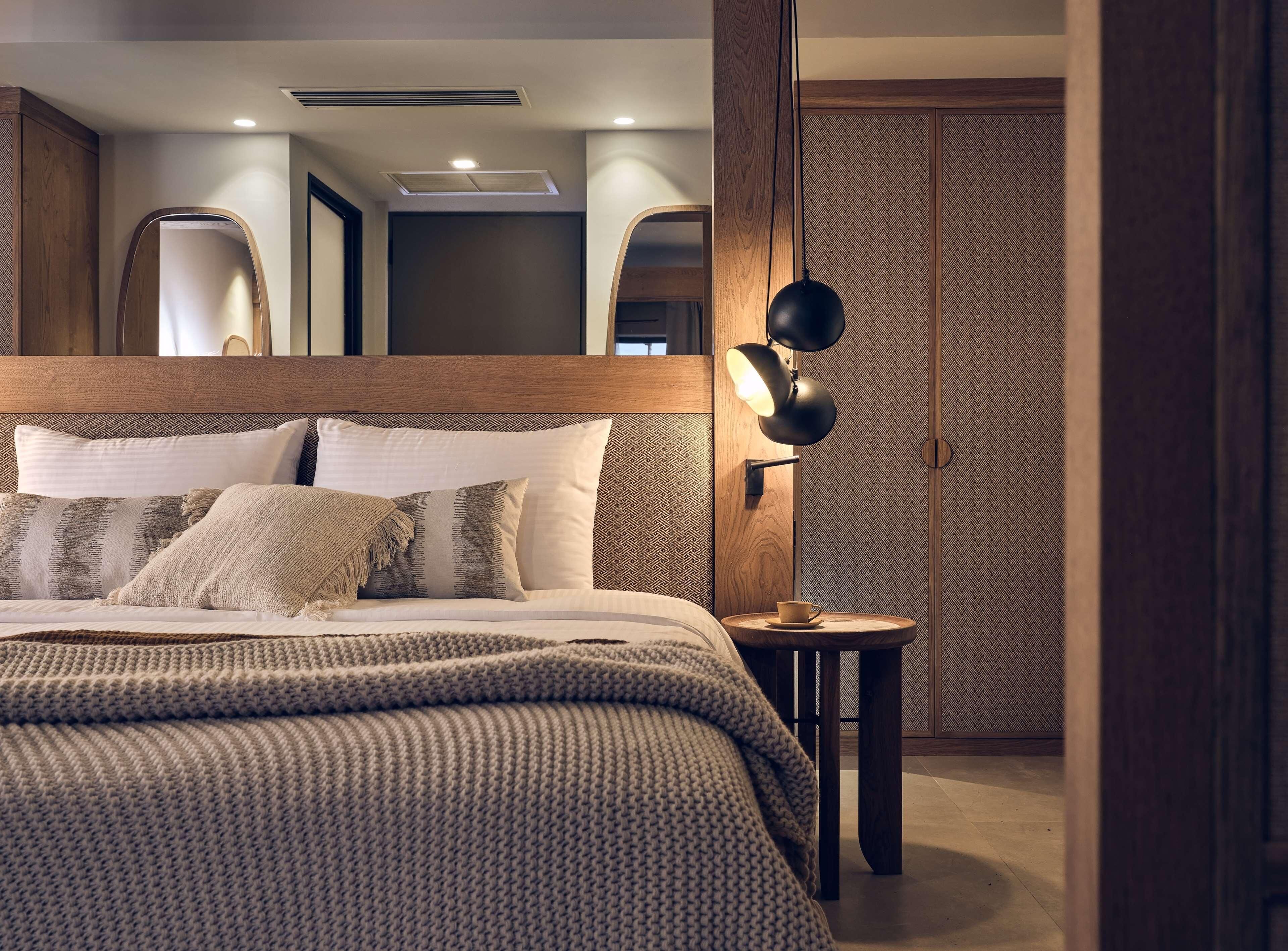 Une chambre à coucher moderne avec une tête de lit en bois, une table de chevet ronde surmontée d’une lampe, et un mur orné d’un motif.