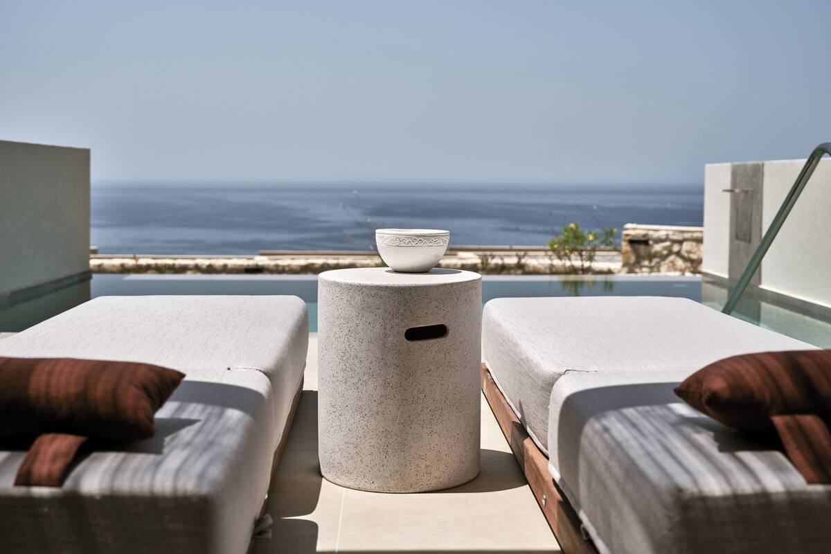 A balcony with two lounge chairs, a small round table, and a bowl. The view includes the ocean and a distant shoreline.
