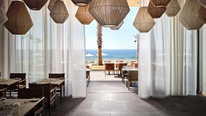 A restaurant with tables and chairs, a view of the beach, and hanging lamps. - The Royal Senses Resort & Spa Crete, Curio Collect (Mylopotamos)