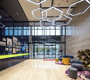 A modern lobby with a wooden floor, hexagonal ceiling lights, a seating area with cushions, and a bookshelf. - Hampton APT Hilton Hangzhou Olympic (HANGZHOU)