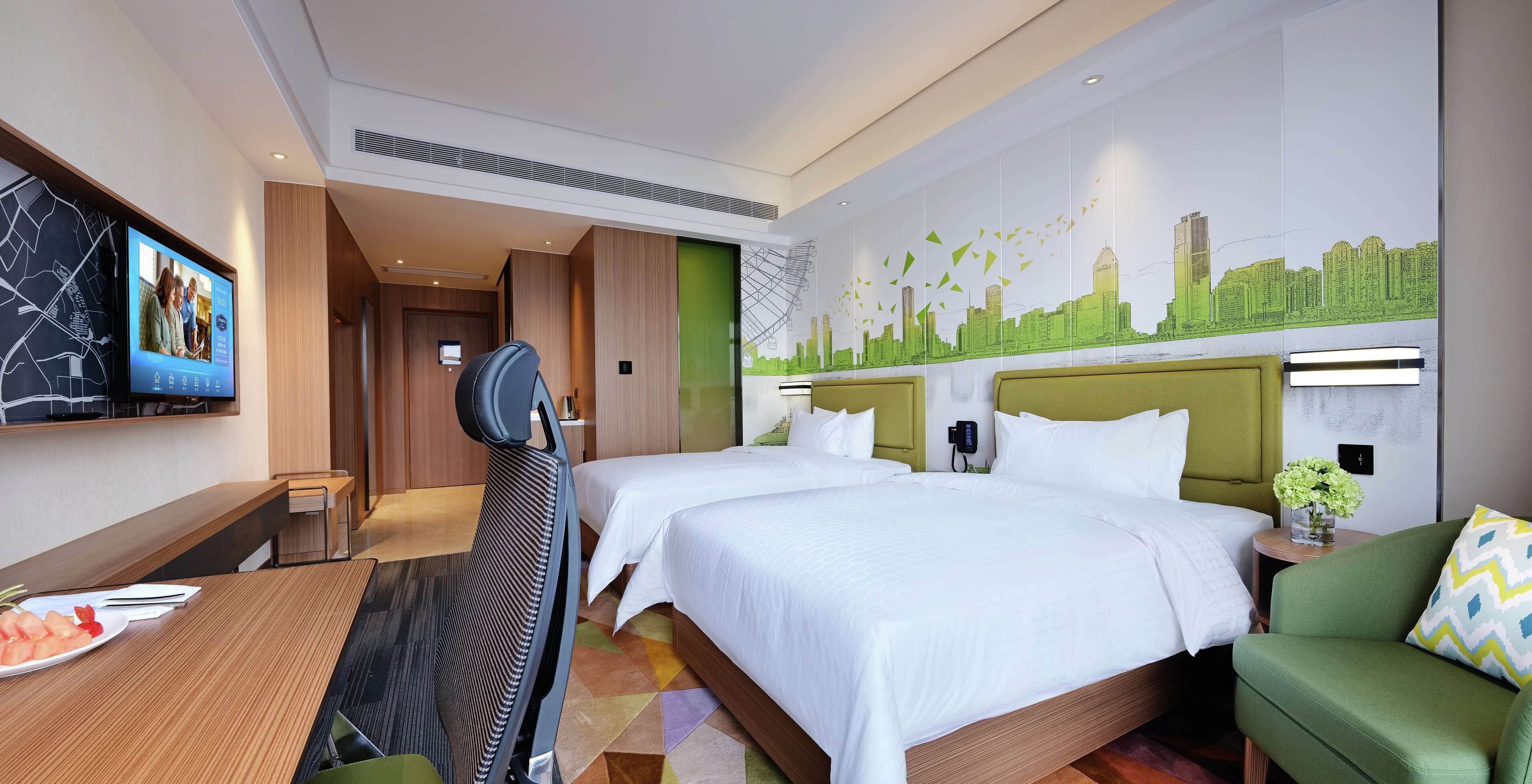 A hotel room with two beds, a desk with a chair, a TV, and a cityscape mural on the wall.