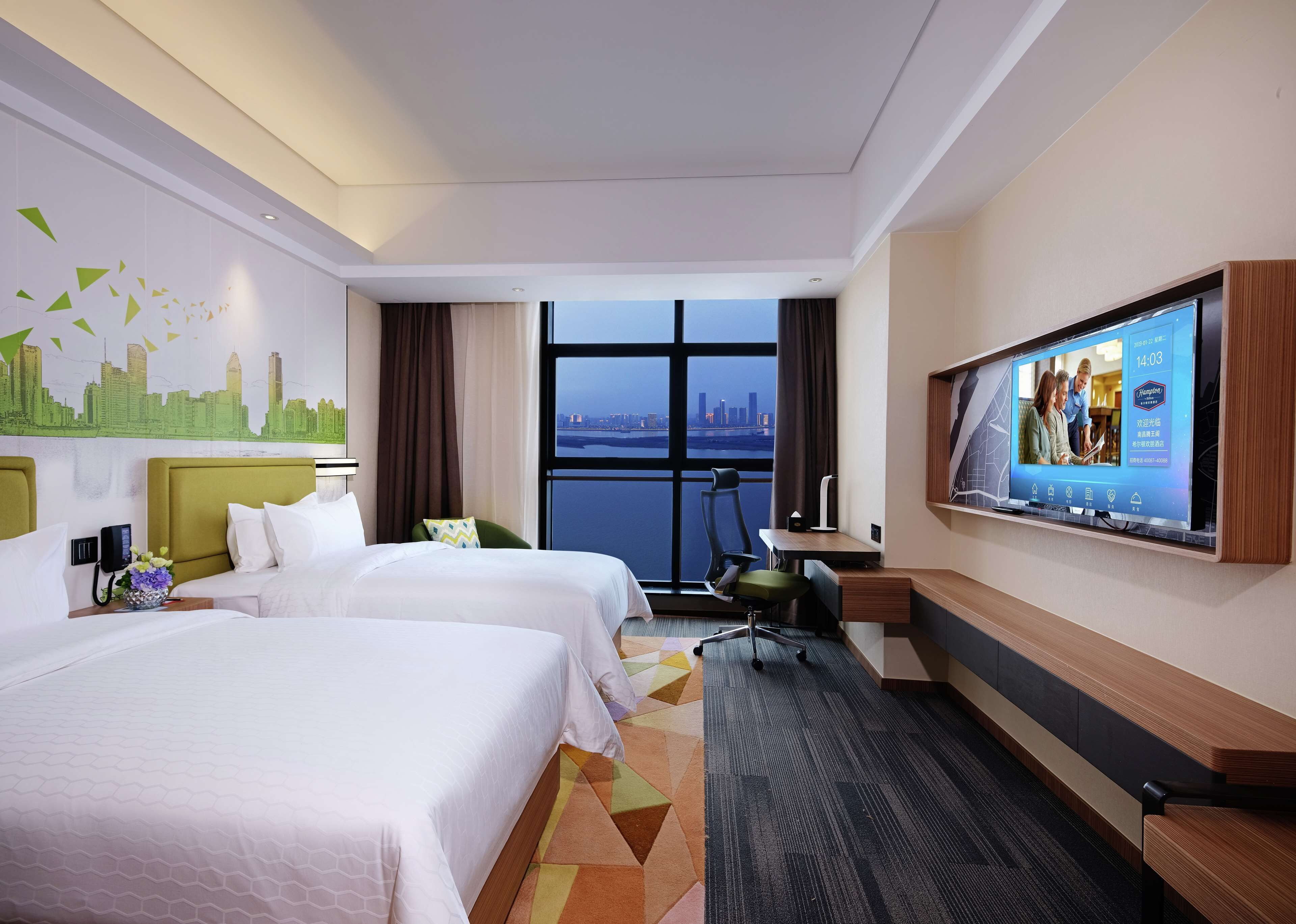 A hotel room with two beds, a desk, a chair, a TV, and a large window with a city view.