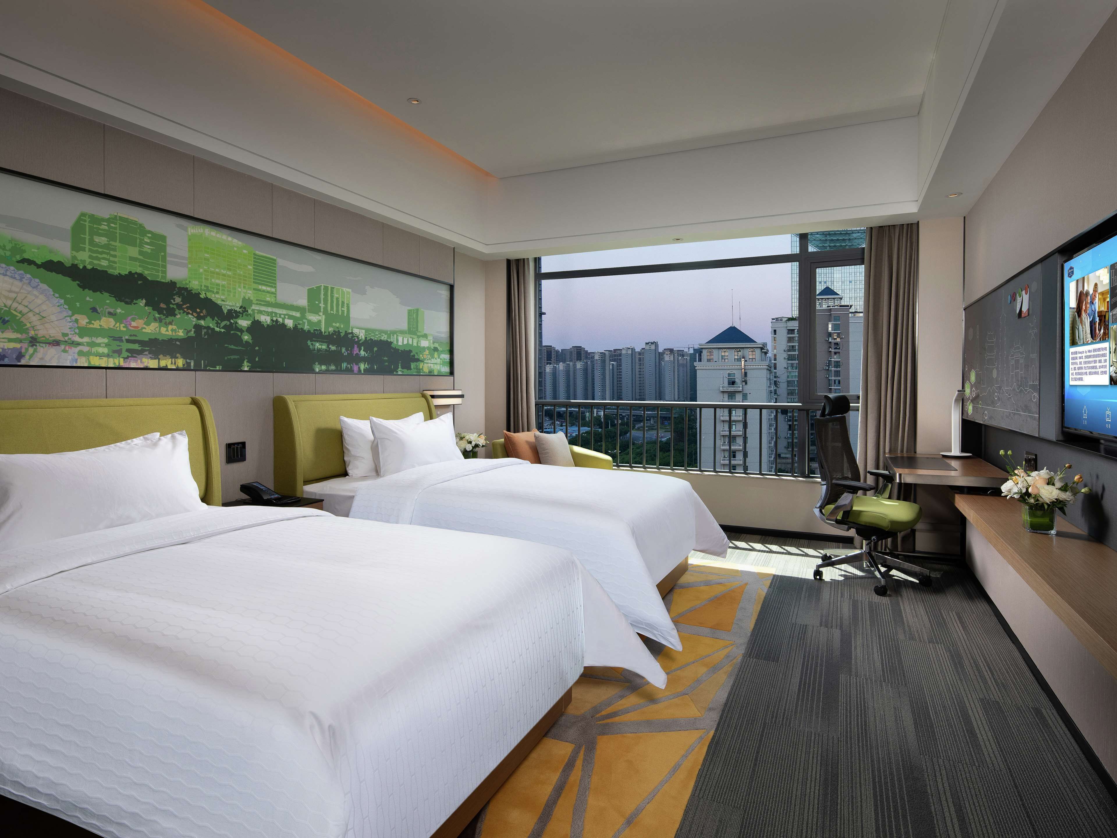 A hotel room with two beds, a desk, a chair, a TV, and a large window with a city view.