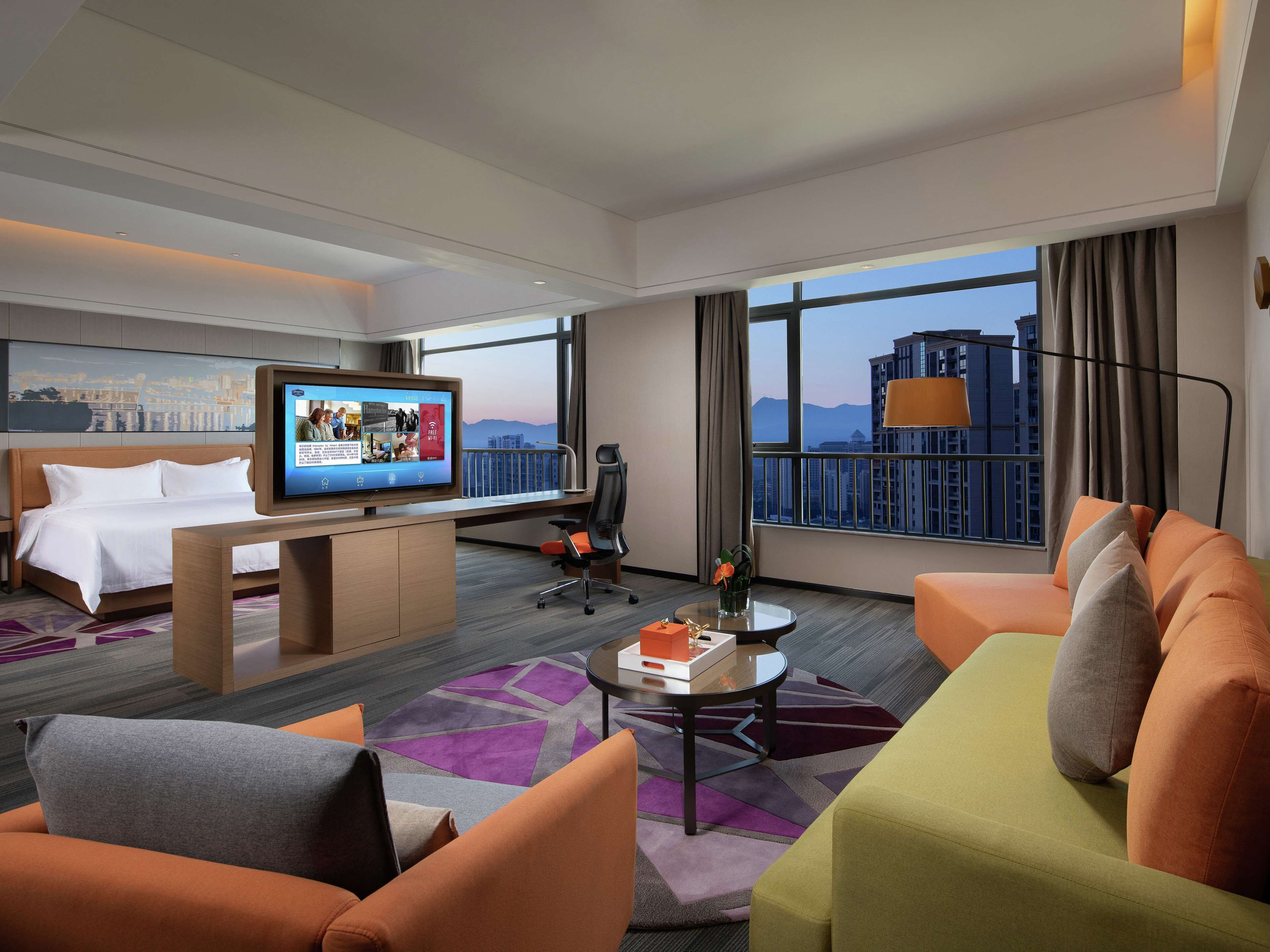 Habitación de hotel con cama, escritorio con televisor, sofá y vista a edificios a través de amplias ventanas.
